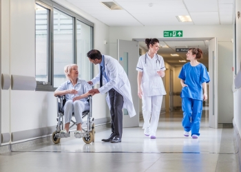 Senior female woman patient in wheelchair sitting in hospital corridor with Asian Indian male doctor and female nurse colleagues