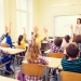 education, elementary school, learning and people concept - group of school kids with teacher sitting in classroom and raising hands
